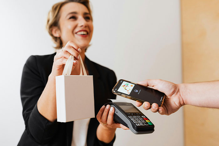 A woman smiles while handing over a small white shopping bag, as a hand holds a smartphone over a card reader for payment.